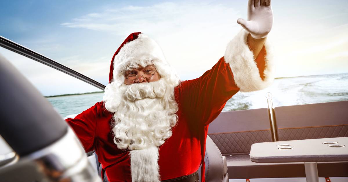A close-up of Santa happily waving while sitting in a boat. The sun is shining over the water behind him. A close-up of Santa happily waving while sitting in a boat. The sun is shining over the water behind him.