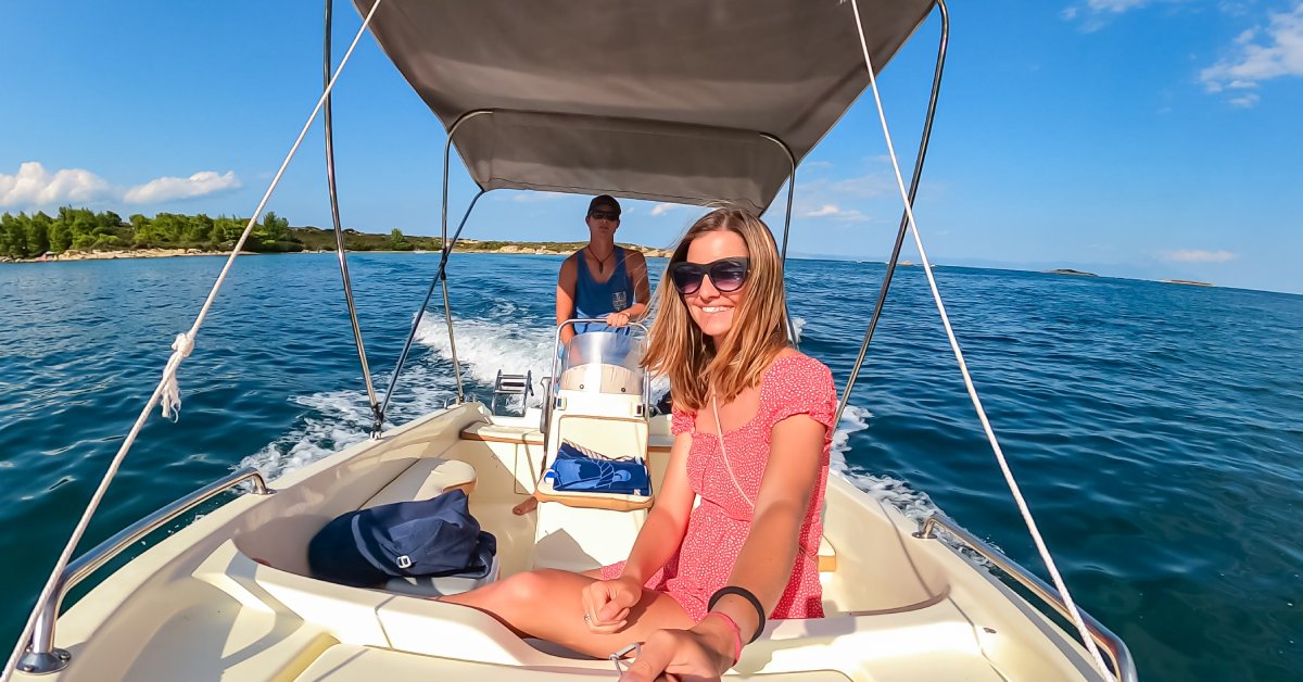 Tips To Make Your First Boat Rental Experience Worth It A man in a blue tank top is driving a boat on a lake. At the front of the boat is a young woman in a pink dress.