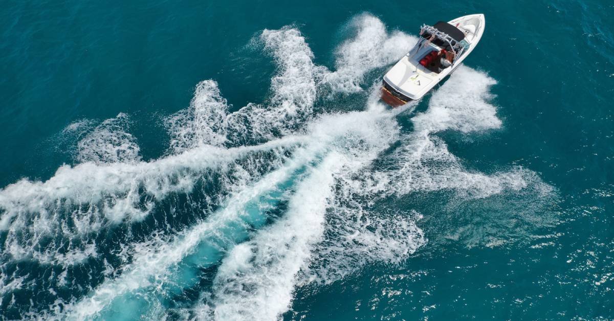 An aerial view of a white boat speeding through the water. A large wake of foaming waves flows behind it. An aerial view of a white boat speeding through the water. A large wake of foaming waves flows behind it.