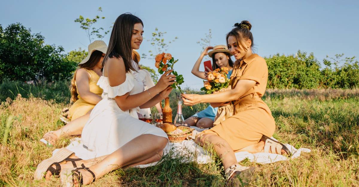Four women in dresses are having a picnic in a lovely field. On the picnic blanket are flowers, bread, and wine glasses. Four women in dresses are having a picnic in a lovely field. On the picnic blanket are flowers, bread, and wine glasses.