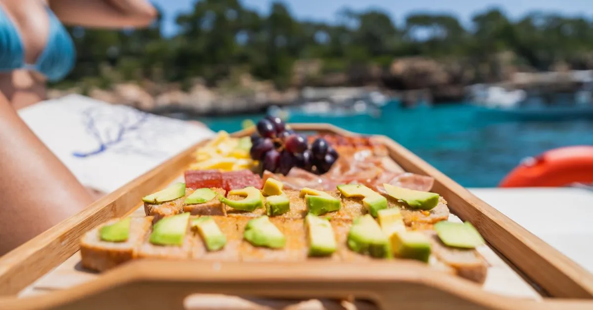 The Best Picnic Recipes for a Summer Day on Your Boat A wooden tray of food is on a boat next to a woman in a bikini. The tray has snacks like avocado toast and fresh grapes.