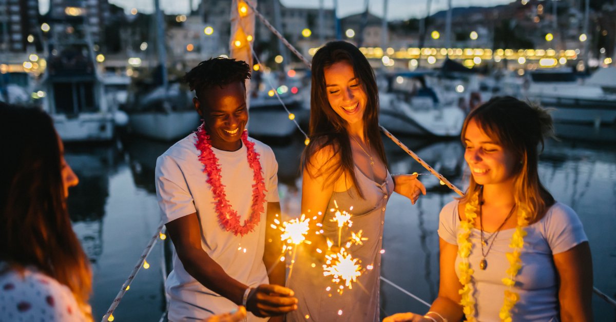 Group of friends holding sparklers at marina during evening, with boats in the background.