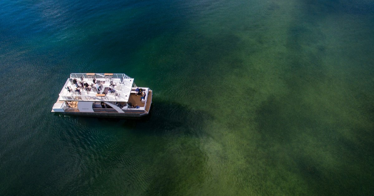 An overhead view of a party pontoon boat on a lake. The pontoon has two levels of seating with numerous people on it.