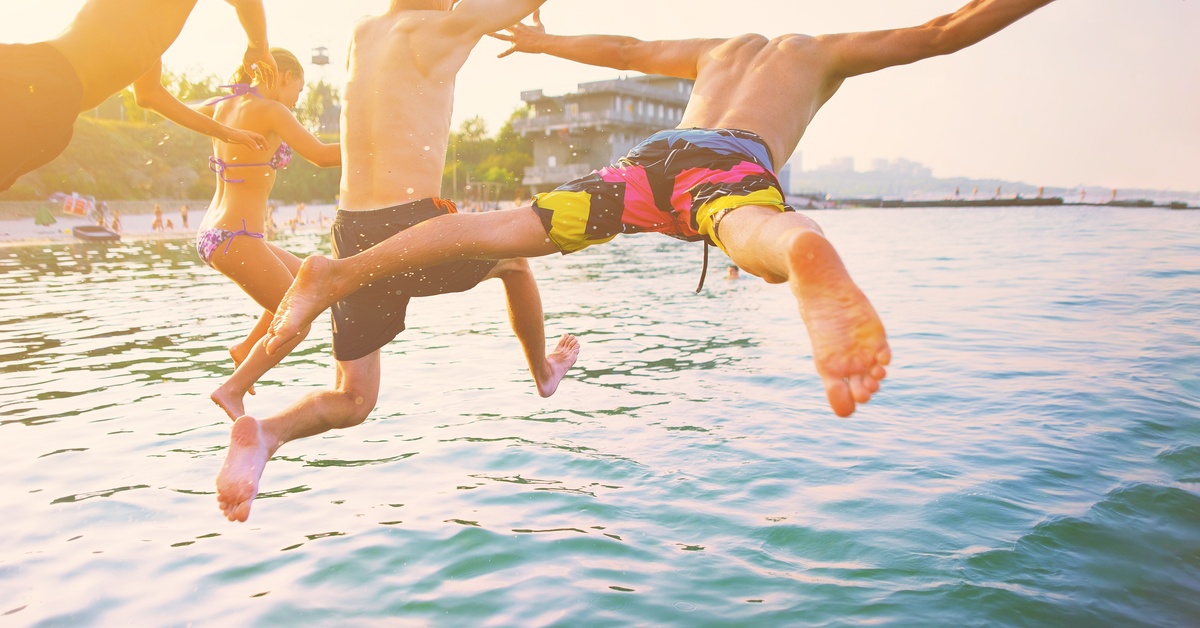 A group of young men and women jumping from a boat into the lake water during the day with a beach in the background.
