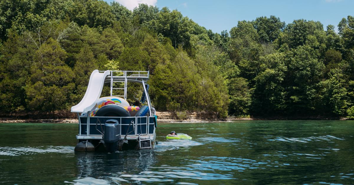 A two-level pontoon with a white slide and multiple inner tubes on a lake during the day, while a woman floats nearby.