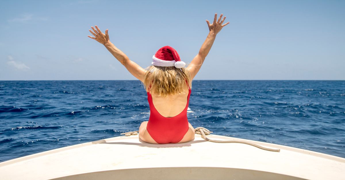 A rear view of a woman in a red one-piece bathing suit with a Santa hat sitting on the bow of a boat with arms raised.
