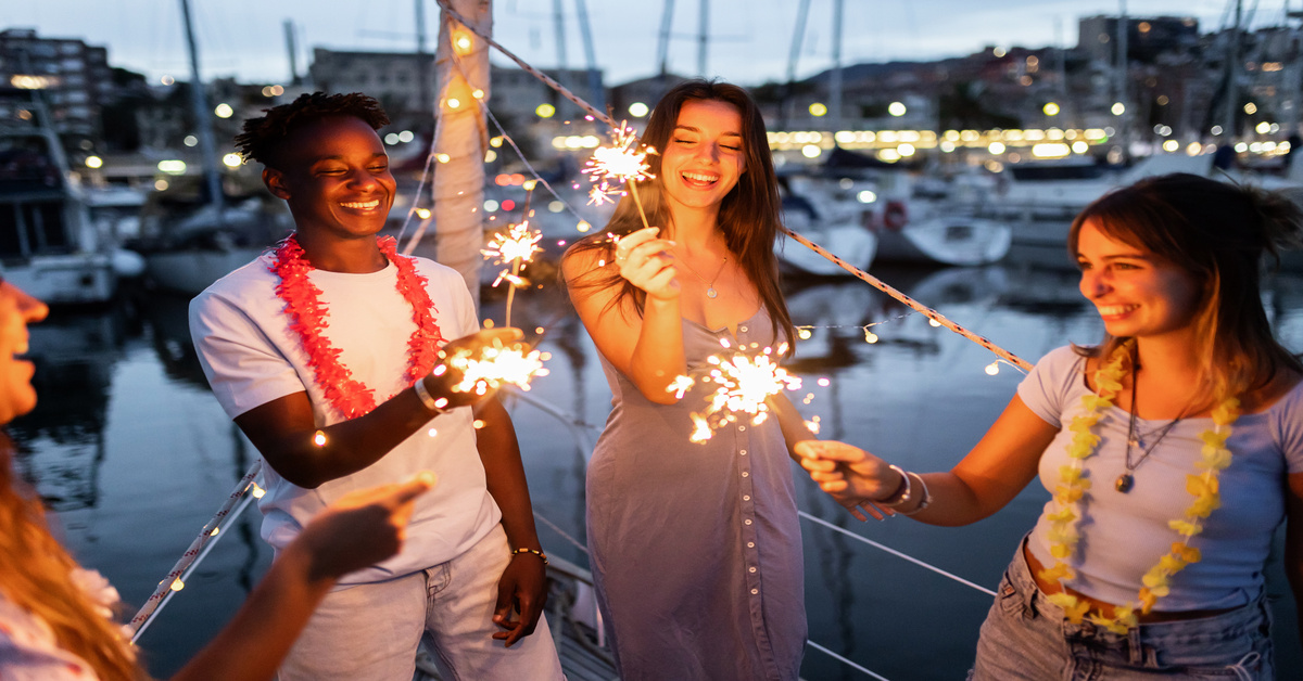 A group of diverse young people on a boat docked in a marina at night, laughing with sparklers in their hands.