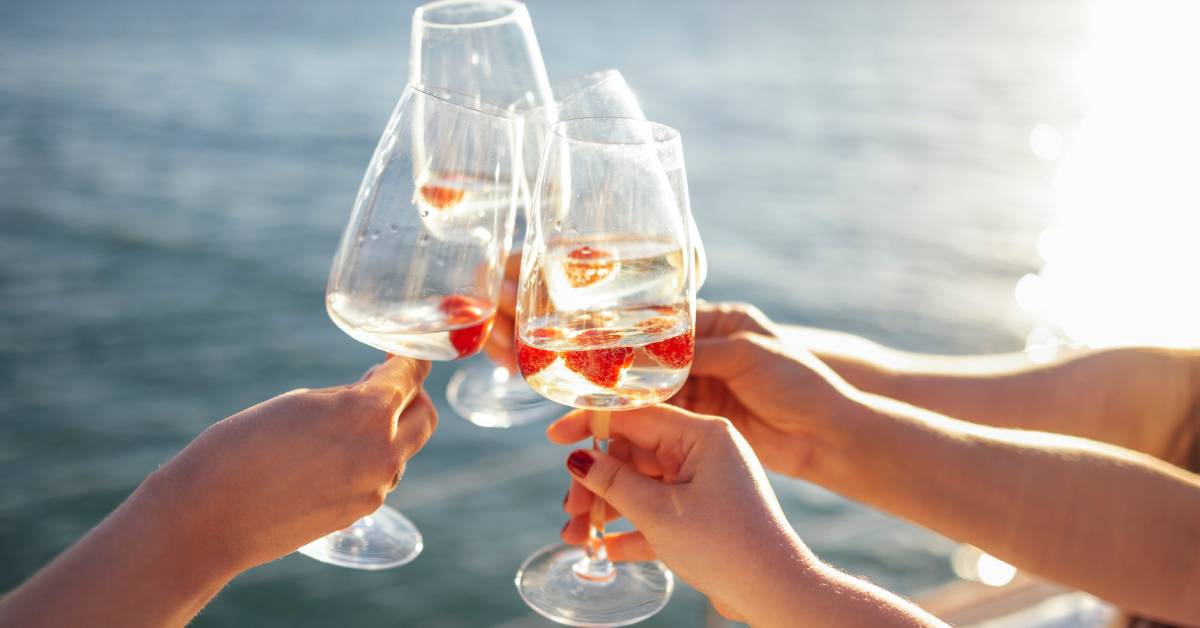 A close-up of a four women toasting their wine glasses with sparkling wine and raspberries on a boat on a sunny day.