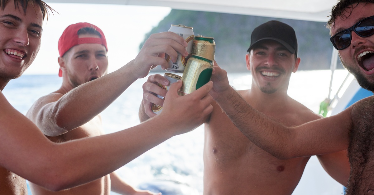 A group of four young men without shirts smiles and cheers as they toast beer cans in the middle of a boat.