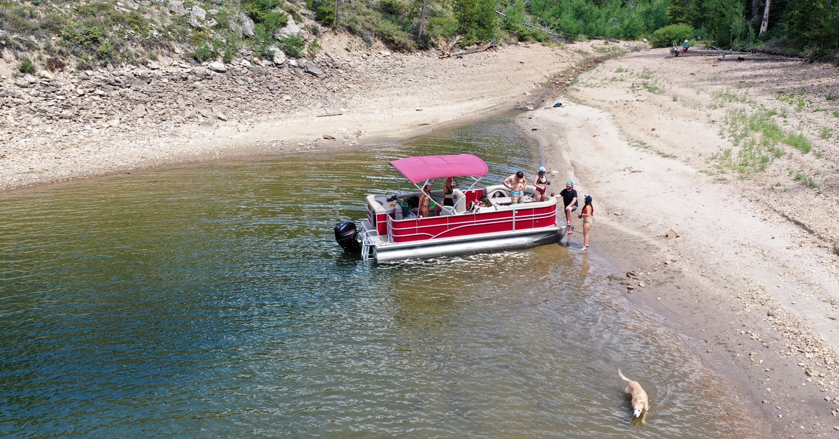A high-altitude view of a red pontoon boat anchored on a lake beach during the day with multiple people aboard.