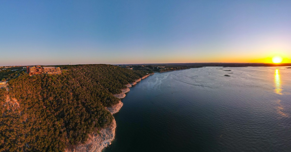 An aerial, panoramic view of Lake travis and the Oasis at sunset, with calm waters, small islands, and surrounding forests.